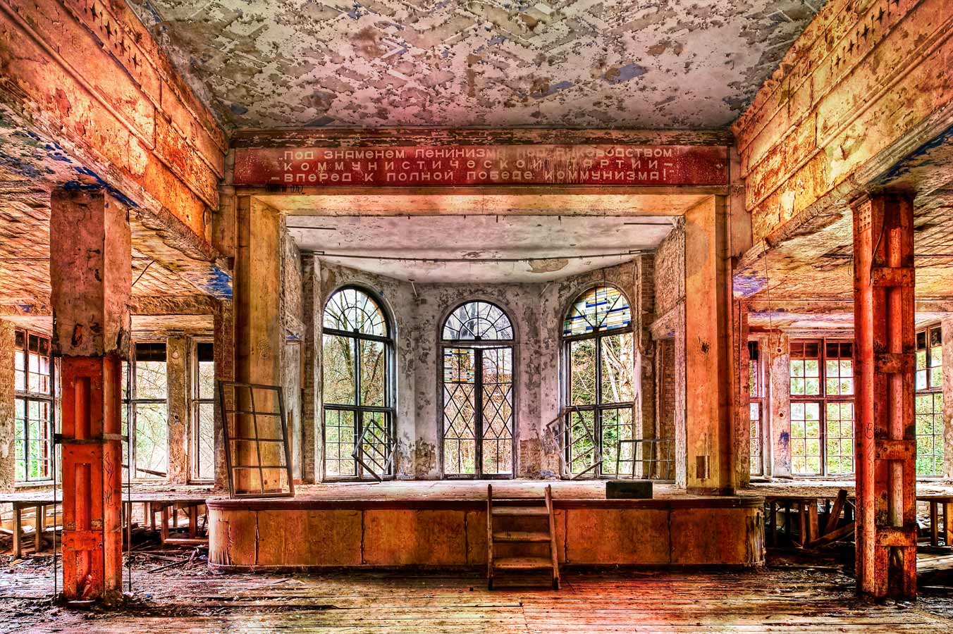 Stage Left

A former hospital, but less well known than Beelitz. Also occupied by the Soviets until the fall of the Wall, I came across this room which must have been intended to entertain the patients. Lots of detail in this image, like the curtain rings above the stage and loudspeaker holes left and right. A Russian speaking colleague translated the text above the stage, it reads: 'Beneath the Flag of Leninism, under the Rulership of the Communist Party - Forward to the Total Victory of Communism'. I spent some time here thinking about the shot and waiting for the wobbly floorboards to settle! I used a shift lens for this image to take in the upper converging lines leading to the stage.