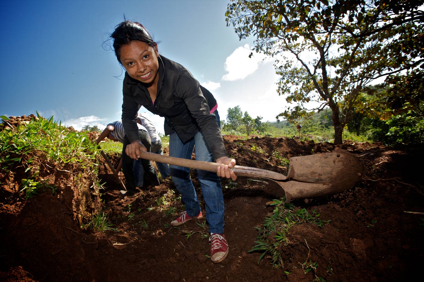 A young woman digs with a shovel, looking for gold ore that has been discarded by the large nearby mine. Low-grade ore that the large mine discards can be processed manually for a modest return.